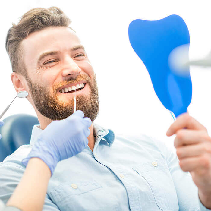 Feel confident in your smile with our porcelain veneers A male patient sitting in a dental chair holding a blue mirror and looking at his teeth