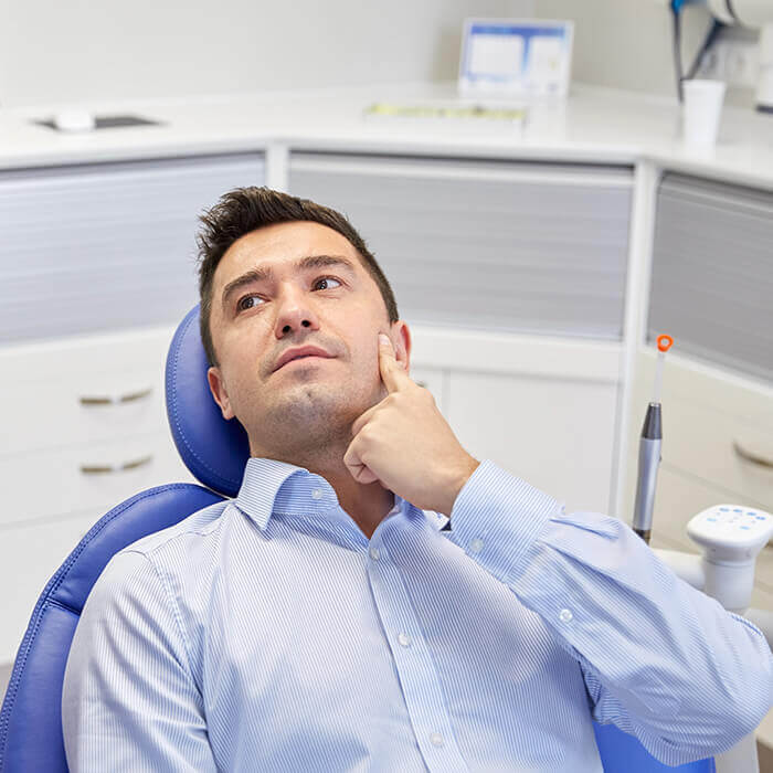 Our team will help you repair teeth damage due to trauma A male patient wearing a blue polo lying in a dental chair pointing at his cheek while looking towards a team member on his side
